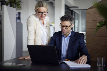 Geschäftsmann, der einen Laptop benutzt und neben einem Kollegen sitzt, der am Schreibtisch im Büro steht - RBF08687