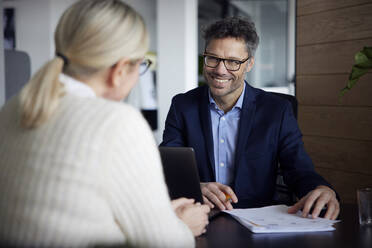Happy businessman discussing ideas with colleague at desk in office - RBF08688