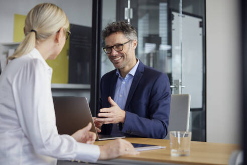 Happy businessman with laptop sitting with colleague at desk in office - RBF08698