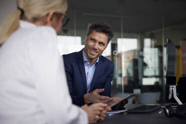 Lächelnder Geschäftsmann, der mit einem Kollegen im Büro über einen Tablet-PC diskutiert - RBF08719