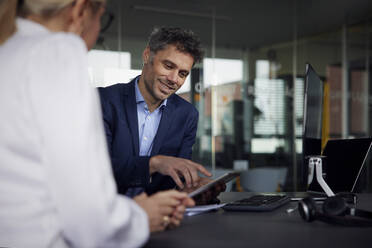 Lächelnder Geschäftsmann, der mit einer Geschäftsfrau im Büro über einen Tablet-PC diskutiert - RBF08720