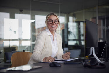 Smiling businesswoman wearing eyeglasses sitting at desk in office - RBF08721