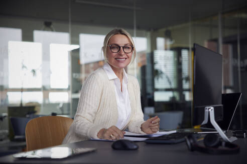 Lächelnde Geschäftsfrau mit Brille sitzt am Schreibtisch im Büro - RBF08721