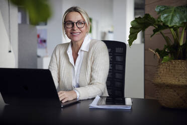 Lächelnde Geschäftsfrau mit Laptop sitzt am Schreibtisch im Büro - RBF08726