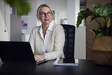 Thoughtful businesswoman sitting with laptop at desk in office - RBF08727