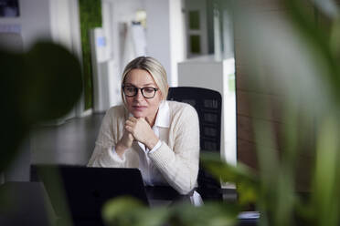 Nachdenkliche Geschäftsfrau mit Händen am Kinn, die auf einen Laptop im Büro schaut - RBF08728