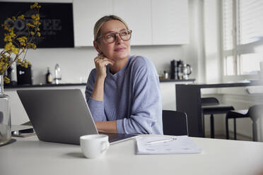 Thoughtful businesswoman wearing eyeglasses sitting with laptop at table - RBF08740