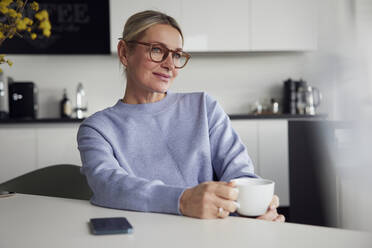 Businesswoman wearing eyeglasses holding coffee cup sitting at table - RBF08743