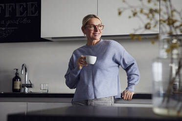 Smiling woman holding coffee cup leaning on kitchen counter - RBF08748