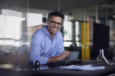 Glücklicher Geschäftsmann mit Brille sitzt am Schreibtisch im Büro - RBF08750