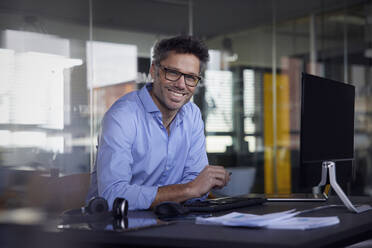 Happy businessman wearing eyeglasses sitting at desk in office - RBF08750