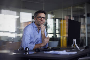 Thoughtful businessman sitting with hand on chin at desk in office - RBF08755