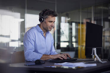 Businessman wearing wireless headset using computer sitting at desk in office - RBF08756