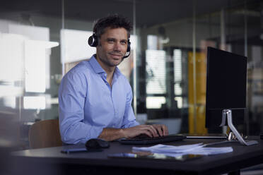 Smiling businessman wearing wireless headset sitting at desk in office - RBF08757