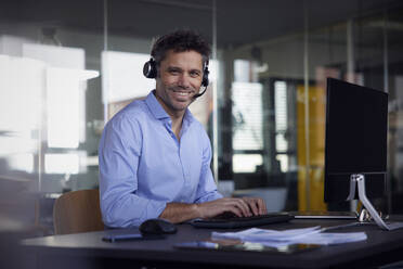 Happy businessman wearing wireless headset sitting at desk in office - RBF08758