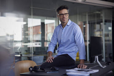 Businessman wearing eyeglasses sitting on desk in office - RBF08762