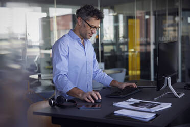 Lächelnder Geschäftsmann mit Brille am Computer stehend am Schreibtisch im Büro - RBF08763