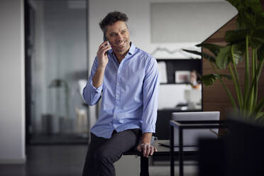 Happy businessman talking on smart phone sitting on desk in office - RBF08773