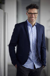 Happy businessman wearing eyeglasses standing with hands in pockets at office - RBF08777