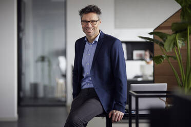 Smiling businessman wearing eyeglasses sitting on desk at work place - RBF08780