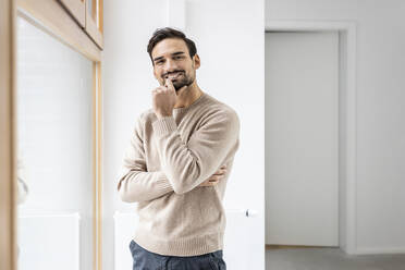 Smiling man with hand on chin standing near window at home - PESF03537