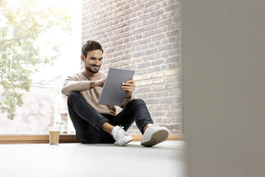 Mann mit Tablet-PC vor einem Glasfenster zu Hause sitzend - PESF03549