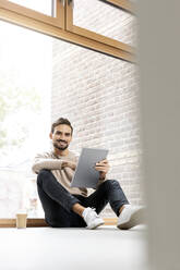 Mann mit Tablet-PC vor einem Glasfenster sitzend - PESF03550