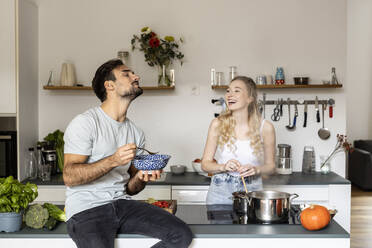 Woman looking at man eating food sitting on kitchen counter at home - PESF03581