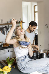 Woman slurping noodles with boyfriend preparing food in kitchen at home - PESF03582