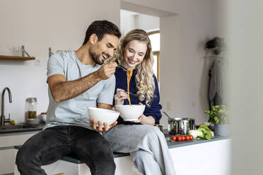 Happy couple eating noodles sitting on kitchen counter at home - PESF03587