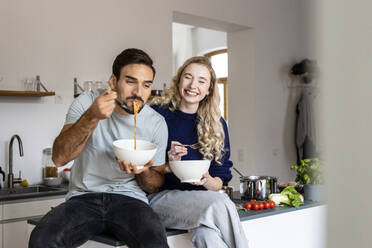 Smiling woman sitting by man eating noodles from bowl in kitchen at home - PESF03588