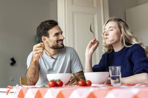 Man looking at girlfriend eating food sitting on dining table at home - PESF03592
