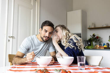 Woman whispering to boyfriend sitting on dining table having food at home - PESF03594