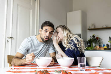 Surprise man listening to girlfriend whispering in ear sitting on dining table at home - PESF03595
