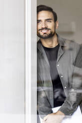 Lächelnder Mann, der sich an die Tür lehnt, gesehen durch Glas - PESF03608