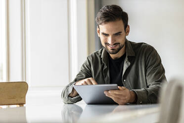 Smiling man sitting at table using tablet PC at home - PESF03614