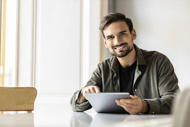 Smiling man sitting at table with tablet PC at home - PESF03615