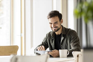 Smiling man using tablet with mug on table at home - PESF03616