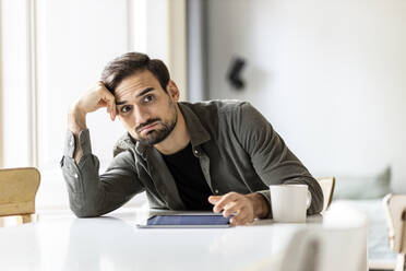 Worried man with tablet PC leaning on table at home - PESF03620