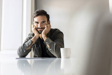 Upset man with tablet PC sitting on table at home - PESF03623