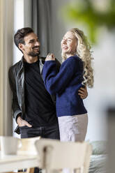 Man with hand in pocket looking at happy girlfriend at home - PESF03629