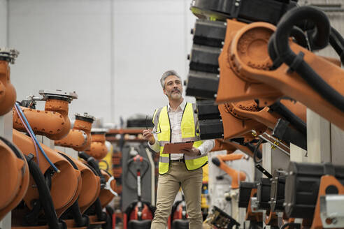 Ingenieur mit Aktenordner und Brille bei der Prüfung von Roboterarmen in einer Fabrik - JCCMF05727