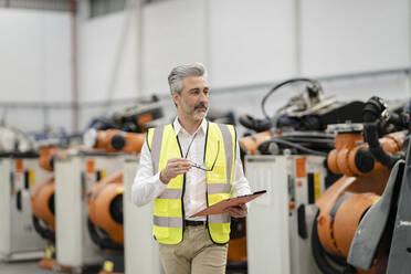 Engineer holding file folder and eyeglasses walking in warehouse at factory - JCCMF05730