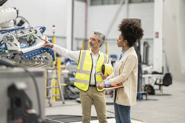 Engineer pointing at robotic arm discussing with businesswoman holding file folder in factory - JCCMF05741