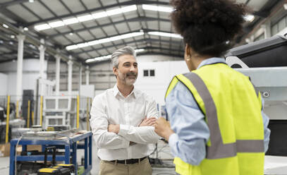 Ingenieur im Gespräch mit einem Geschäftsmann, der mit verschränkten Armen in einer Fabrik steht - JCCMF05766