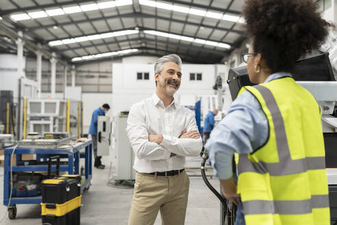 Lächelnder Geschäftsmann mit verschränkten Armen, der mit einem Ingenieur in einer Fabrik diskutiert - JCCMF05769