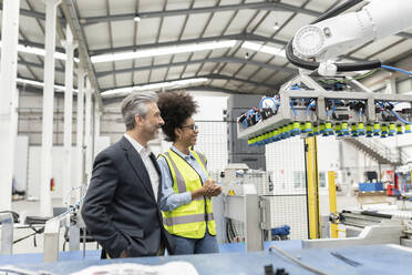 Happy businessman with hands in pockets standing by engineer looking at robotic machine in factory - JCCMF05776
