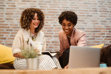 Lächelnde multirassische Kolleginnen in eleganter Kleidung surfen auf Netbooks, während sie gemeinsam an einem Projekt auf einer Couch in einem hellen, modernen Büro arbeiten - ADSF34078