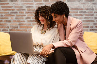 Positiv gestimmte, multirassische Kolleginnen in eleganter Kleidung surfen auf Netbooks, während sie gemeinsam an einem Projekt auf der Couch in einem hellen, modernen Büro arbeiten - ADSF34079