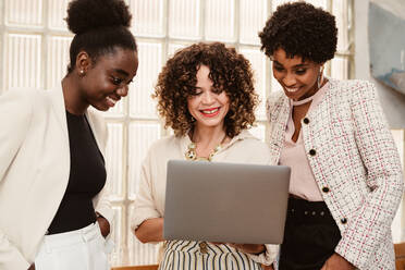Gruppe glücklicher multiethnischer weiblicher Mitarbeiter in eleganter Kleidung beim Surfen auf einem Netbook, während sie gemeinsam an einem Projekt in einem hellen, modernen Büro arbeiten - ADSF34080